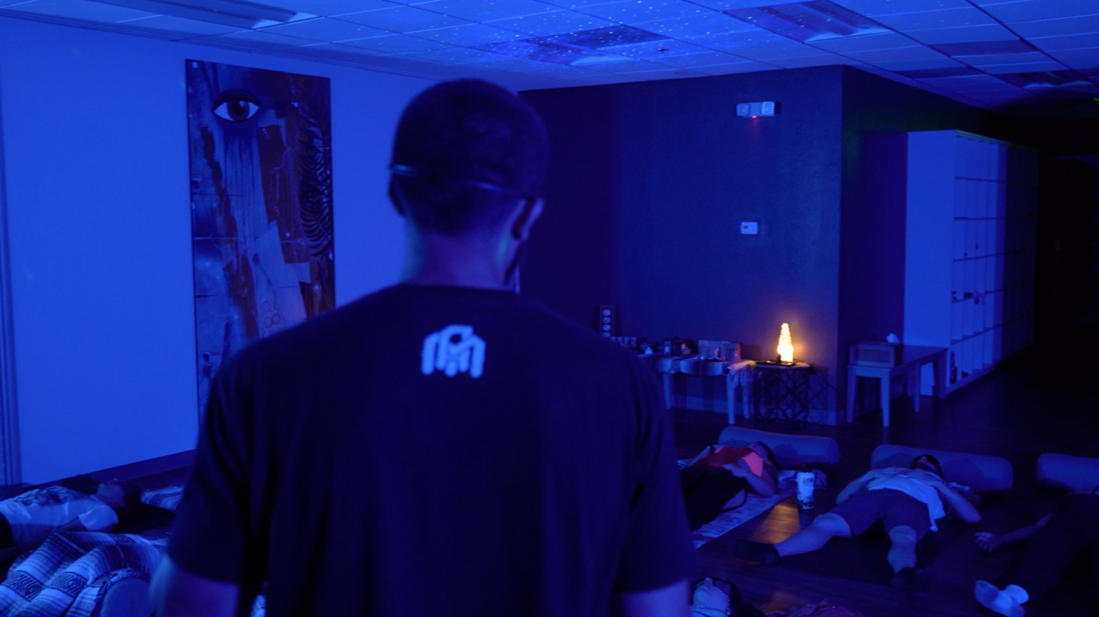 Atmospheric blue-lit live Breathwork room with participants lying down on mats while the facilitator stands at the front, the felt-in-the-body experience that 9D Breathwork tracks try to recreate through headphones